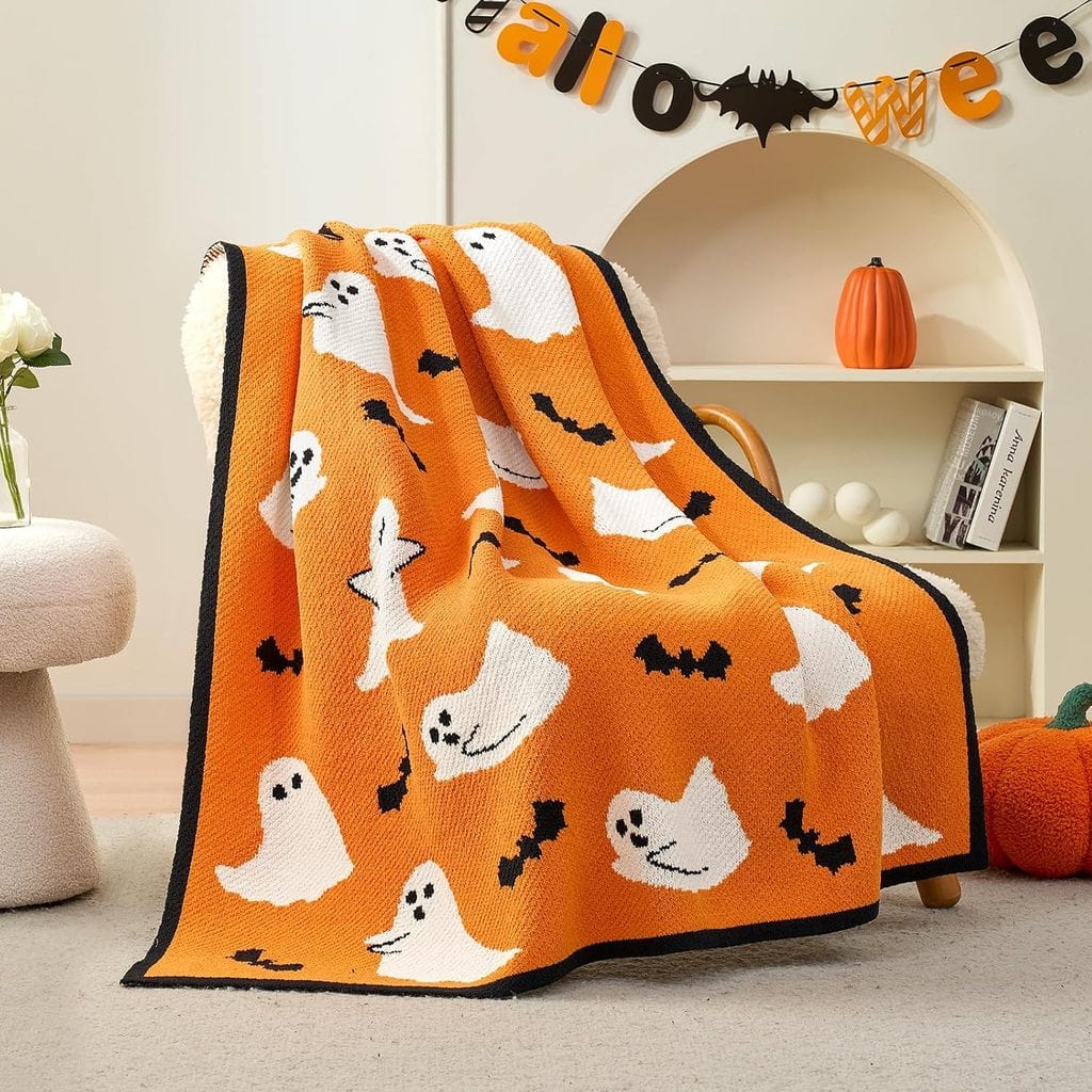 An orange blanket with white ghosts and black bats draped over a chair, with Halloween decorations, a pumpkin, and a “Halloween” banner in the background.