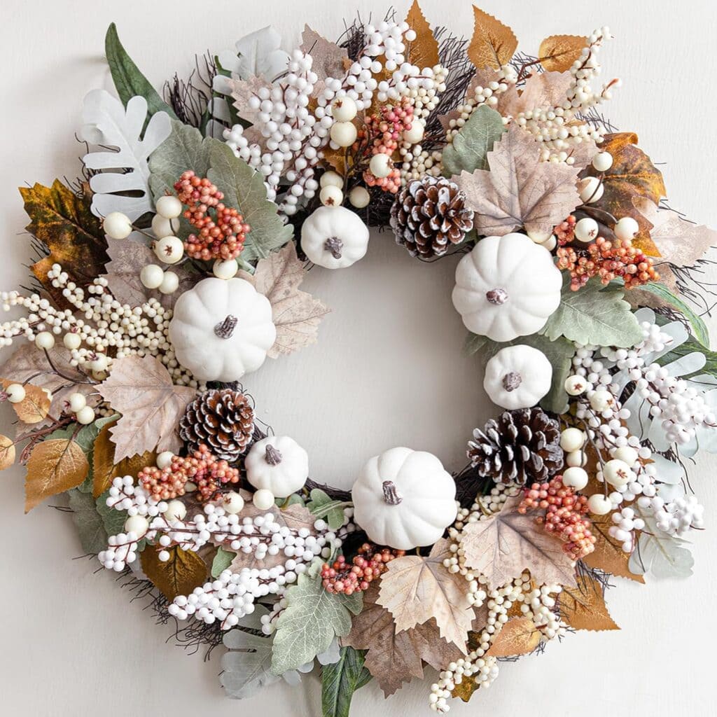 A decorative wreath featuring white pumpkins, pinecones, white berries, pink berries, and assorted autumn leaves in muted tones, arranged in a circle on a light background.