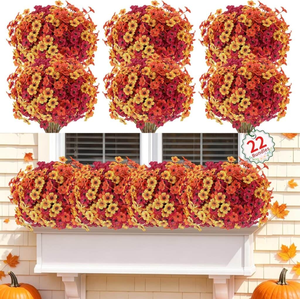 Twenty-two bundles of artificial orange, red, and yellow flowers are arranged in a window box on a white house, with autumn leaves scattered around.