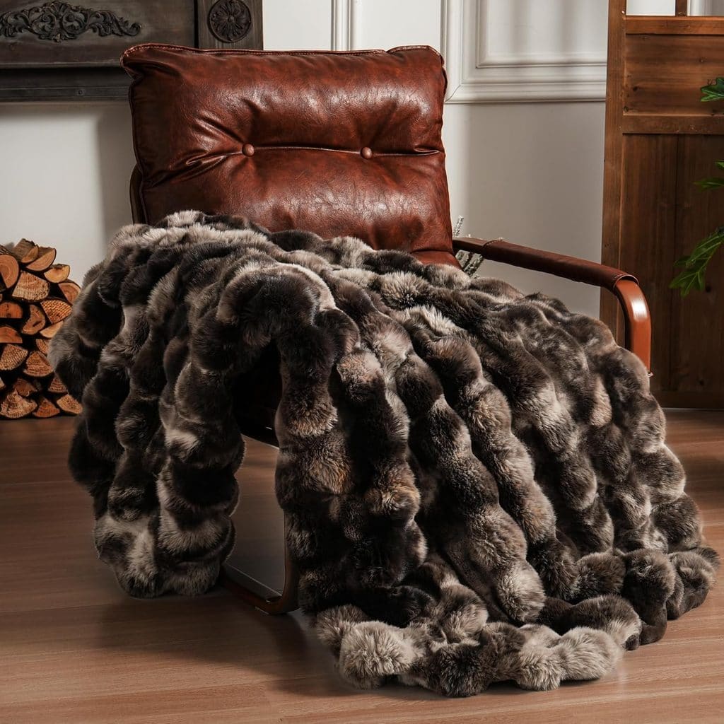 A plush faux fur blanket with shades of brown, gray, and cream is draped over a brown leather armchair in a cozy room with wood floors and a stack of firewood in the background—perfect for relaxing evenings, especially when you pick it up during our Thursday Deals.