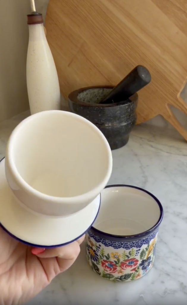 A hand holds a white cup and saucer above a colorful mug on a marble countertop, with a cutting board, mortar and pestle, and a bottle in the background.