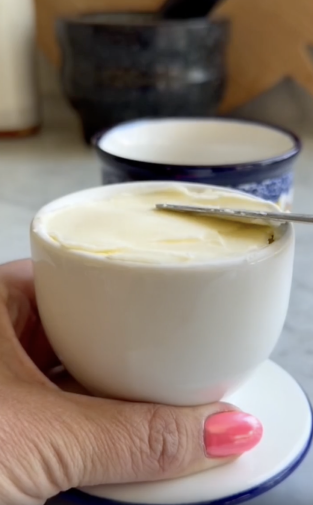 A hand with pink nail polish holds a small white cup filled with creamy butter, with a knife spreading the top. Another cup is blurred in the background.