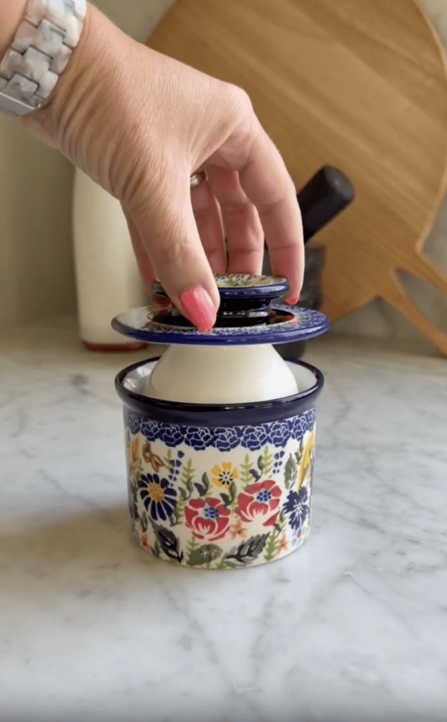 A hand with pink nails lifts the lid off a colorful, floral-patterned ceramic container on a marble countertop, with a wooden cutting board in the background.