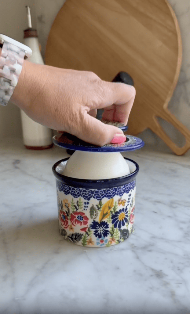 A hand lifts the blue floral lid off a matching ceramic butter dish on a marble countertop; a wooden cutting board and bottle are in the background.