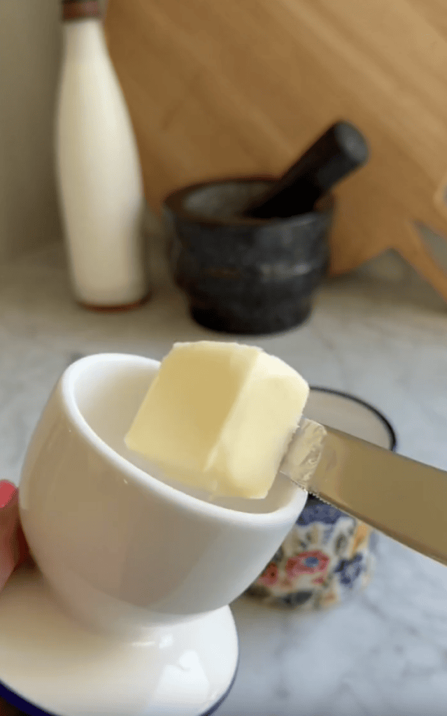 A knife holds a cube of butter over a white egg cup on a kitchen countertop, with a mortar and pestle and a bottle in the blurred background.