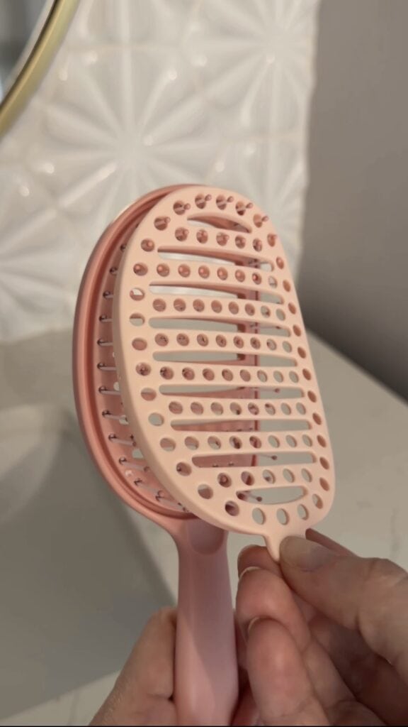 A close-up of a light pink hairbrush with a slotted design and bristles, held in a hand. The background shows a white textured wall and a light-colored surface.