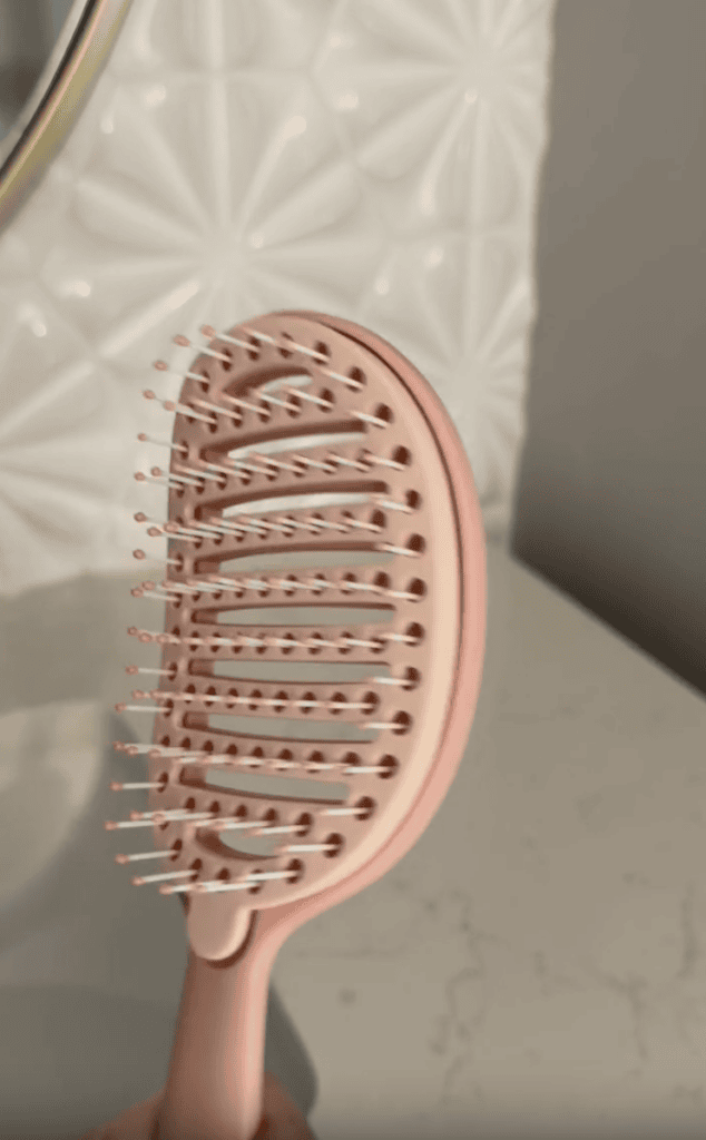 A close-up of a pink vented hairbrush with white bristles, held in front of a textured white tile wall and a light-colored countertop.
