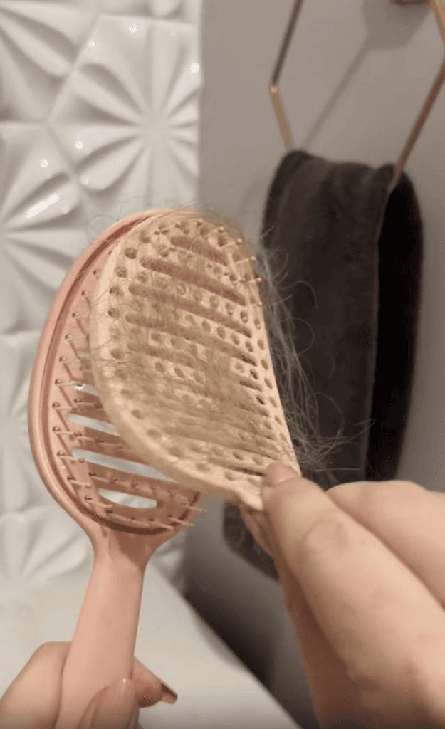 A hand removes light brown hair from a pink hairbrush with a flexible, vented design. A dark towel hangs on a gold holder against a white textured bathroom wall in the background.