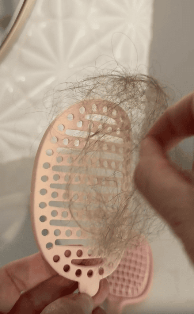 A hand removing a clump of hair from a pink brush cleaning tool with holes, against a white textured background.