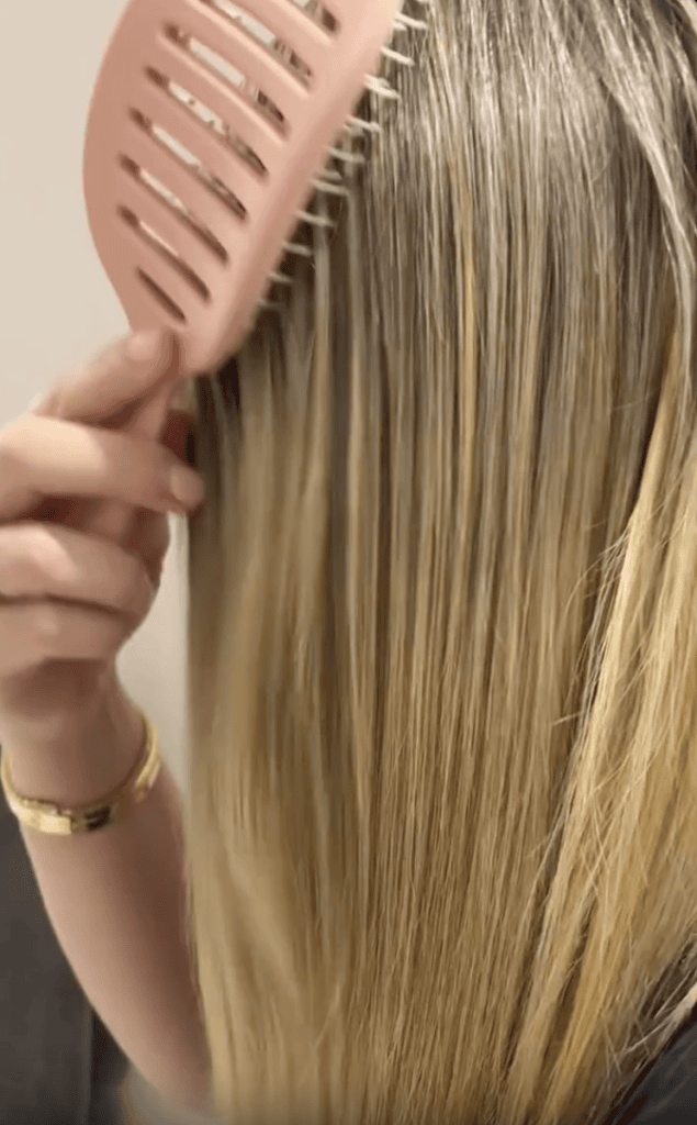 A person with straight, blonde hair is brushing their hair with a wide-tooth pink comb. They are wearing a gold bracelet on their wrist.