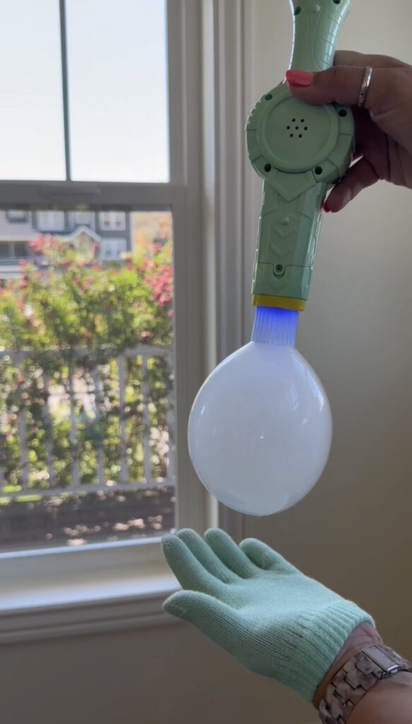 A hand holds a bubble-blowing toy releasing a large bubble, while another hand in a green glove prepares to catch it near a sunlit window with a garden view.