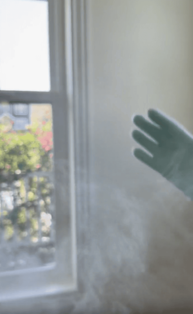 A gloved hand reaches into a sunlit room with a window, with mist or spray visible in the air and greenery seen outside.