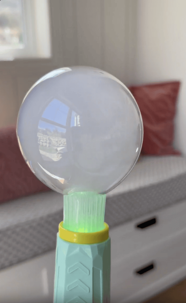 A large soap bubble rests on a green bubble wand, with a window and a white bed with pink pillows in the background.