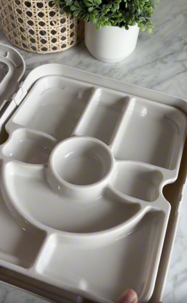 A close-up of an empty, white, divided lunch tray with multiple sections and a round compartment in the center. The tray is held over a marble countertop with potted plants in the background.