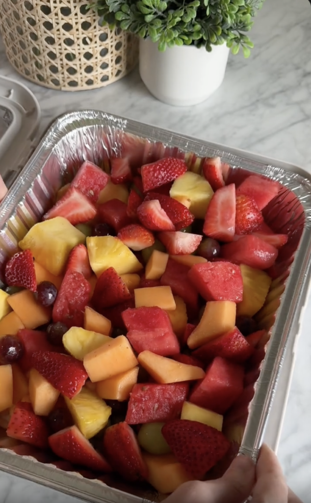 A foil tray filled with freshly cut fruit, including strawberries, watermelon, cantaloupe, pineapple, and grapes, sits on a marble countertop.
