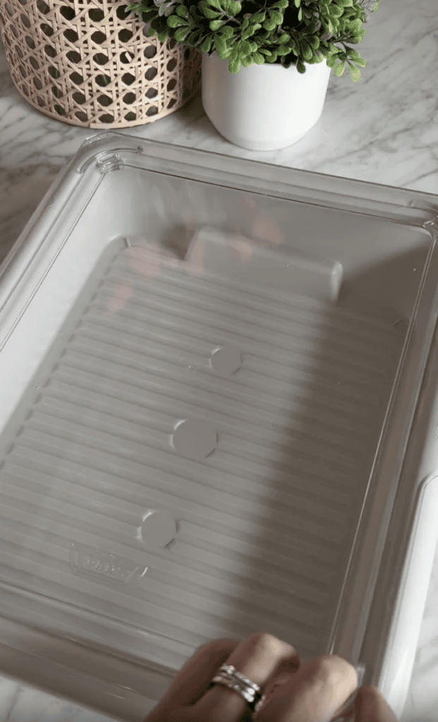 A hand holding a clear plastic container with a ridged bottom, placed on a marble countertop. Green plants and a woven basket are visible in the background.