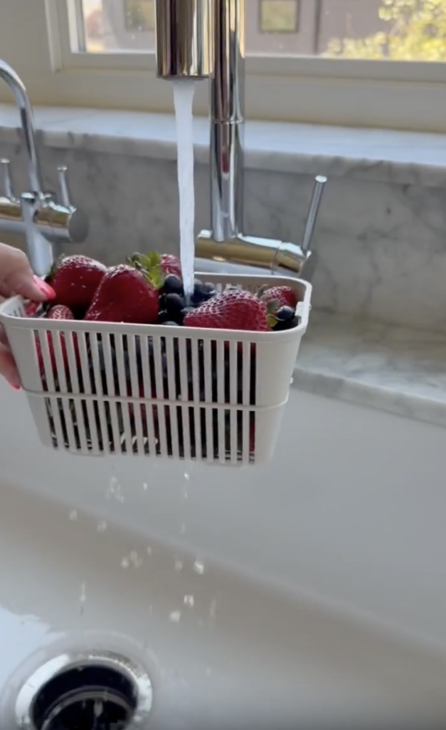 A hand holds a white basket filled with strawberries and blueberries under a running faucet to rinse the berries in a kitchen sink.