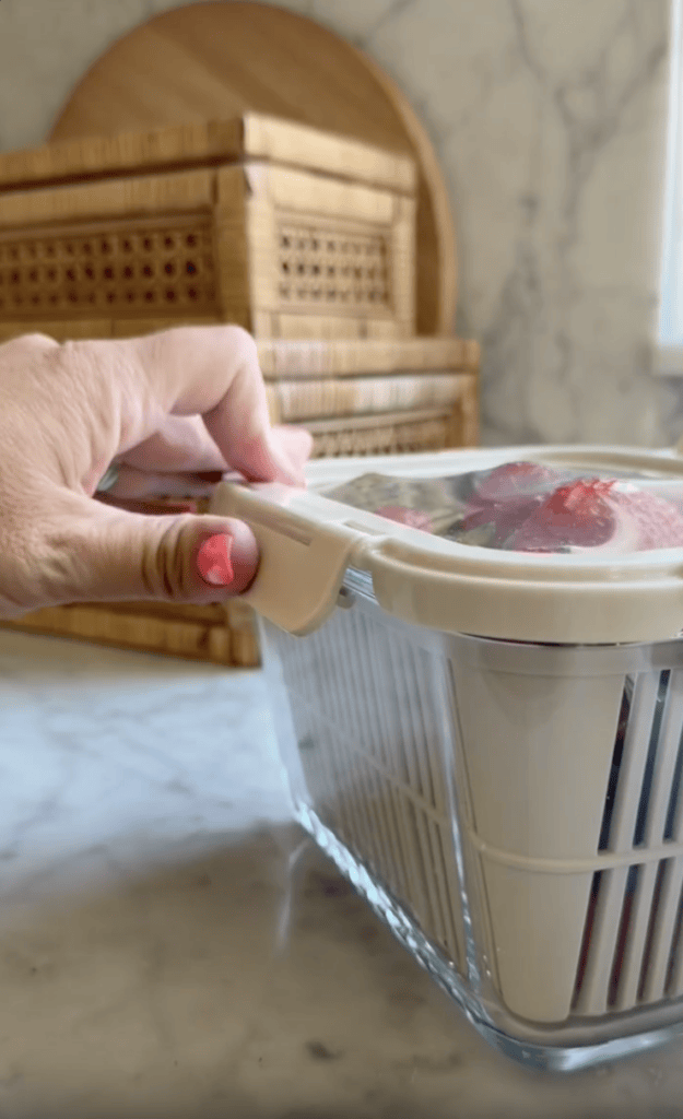 A hand with pink nail polish secures the lid on a glass container filled with strawberries, on a marble countertop with wicker baskets in the background.
