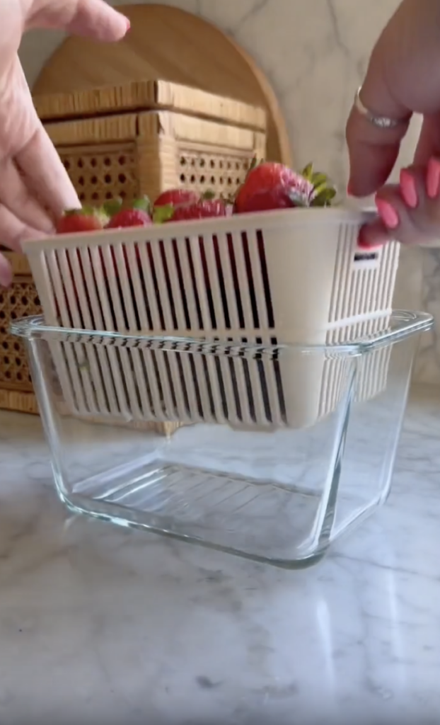 Hands holding a white plastic basket filled with strawberries above a clear glass container on a marble countertop, with wicker items and a cutting board in the background.