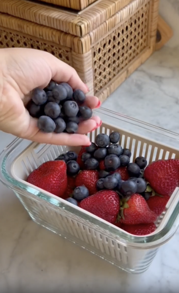 A hand holds fresh blueberries above a glass container filled with strawberries and more blueberries, sitting on a marble countertop near wicker baskets.