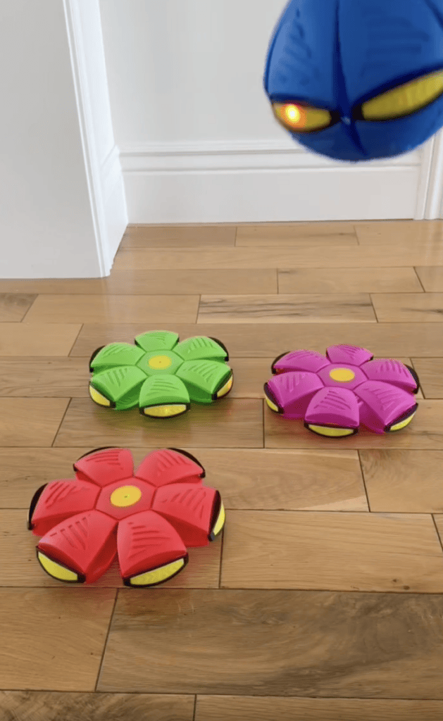 Three flower-shaped toy discs, one green and two pink, are on a wooden floor. A blue disc is suspended above, showing a small orange light. White walls and a door frame are visible in the background.
