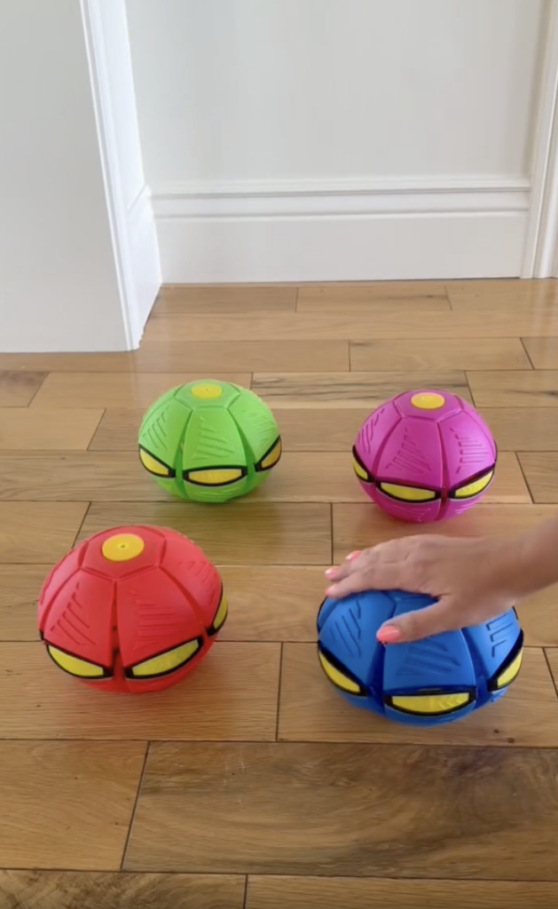 Four colorful, segmented ball-like toys sit on a wooden floor. A hand presses on the blue one, while the others are red and green. A white wall and baseboard are in the background.
