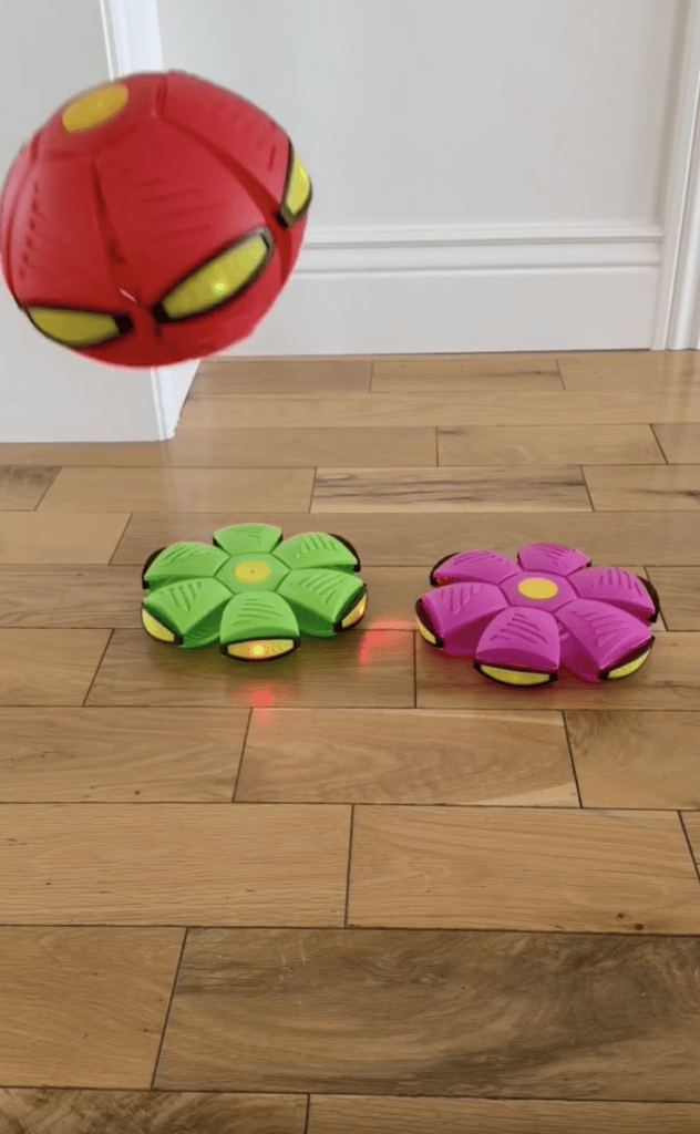 A red disc-shaped toy is in mid-air above a wooden floor, while green and pink flower-shaped disc toys rest on the floor below.