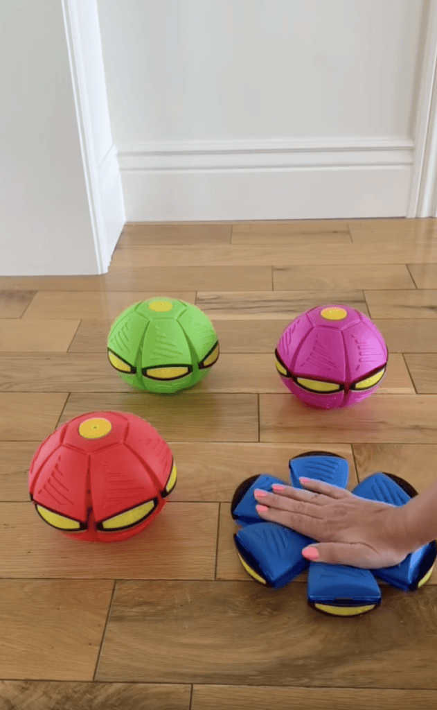A hand presses a blue collapsible ball flat on a wooden floor, next to red, green, and pink similar balls that are fully expanded.