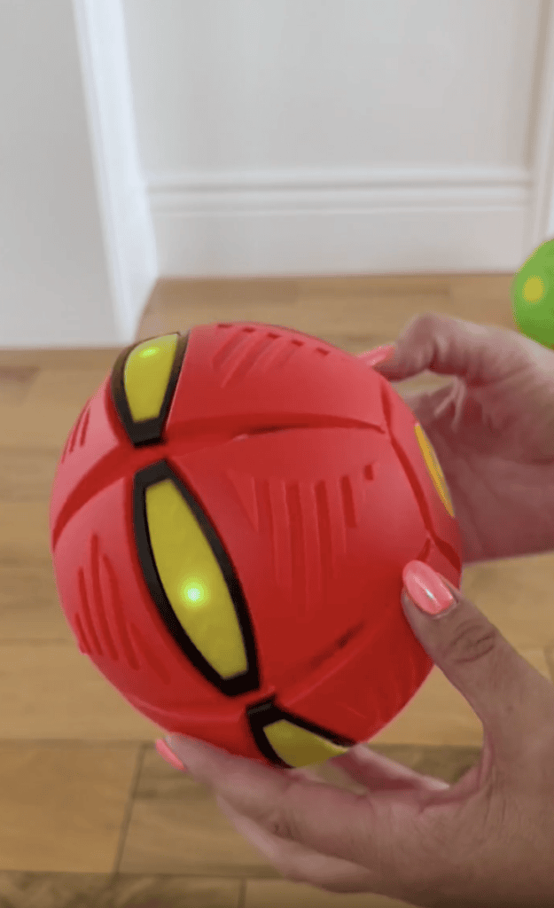 A person holds a red and yellow toy with black lines and a green light, standing on a wooden floor near a white wall.