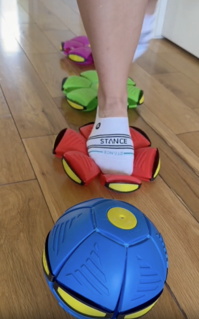 A person wearing white socks steps on collapsing disc toys shaped like flowers on a wooden floor, with a blue ball-shaped toy in the foreground.