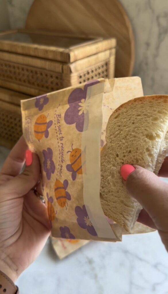 Two hands with pink nails hold a slice of bread and a beeswax food wrap decorated with bees and purple flowers. Wicker boxes are blurred in the background.
