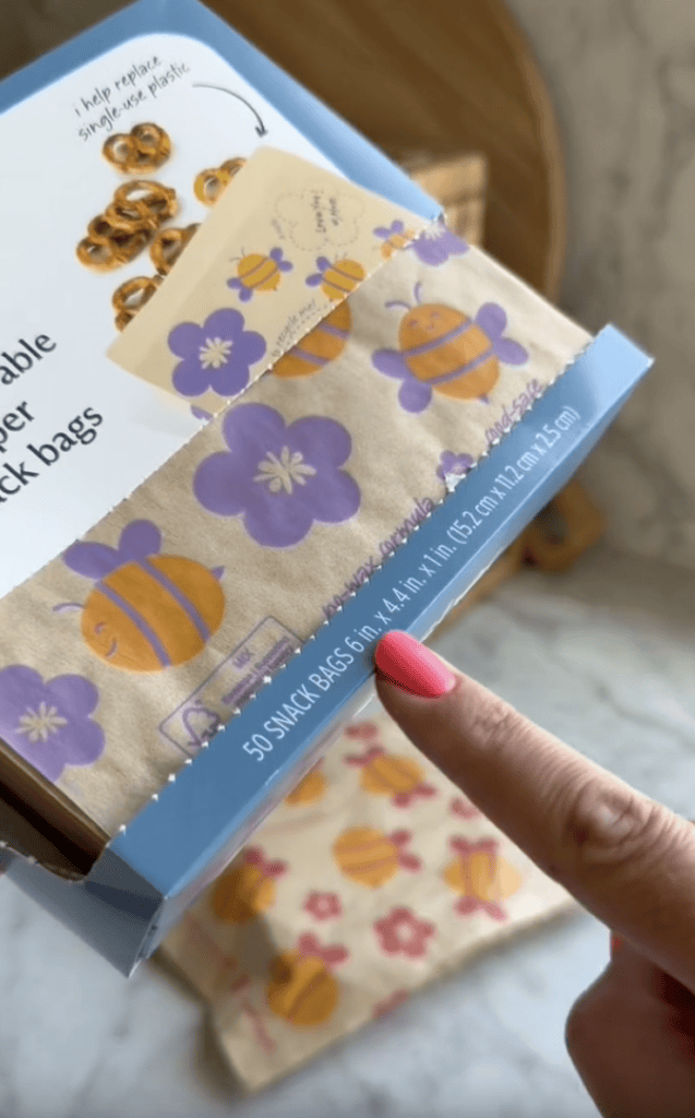 A hand with pink nail polish points at a box of reusable paper snack bags decorated with bees and purple flowers. The box label shows the size: 6in x 4.4in. An open bag is partially pulled from the box.