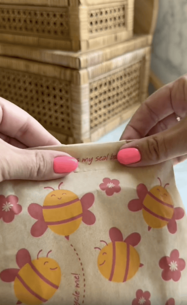 Two hands with pink nail polish hold a brown paper bag decorated with smiling cartoon bees and pink flowers. Woven baskets are visible in the background.