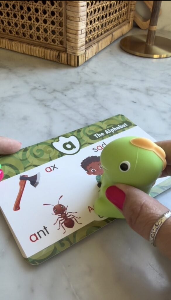 A hand holds a green frog-shaped pointer over a flashcard showing the letter “a,” with images of an axe, an ant, and a cartoon face. Another hand holds the card on a marble surface.