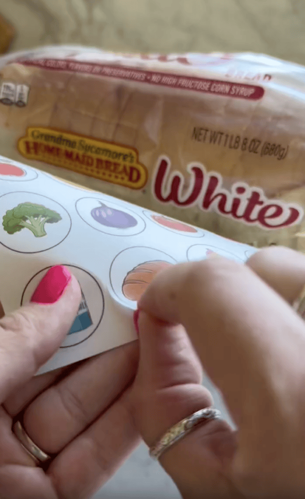 A person with pink nails peels a round vegetable sticker, with a loaf of white bread labeled “Home Maid Bread” in the background.