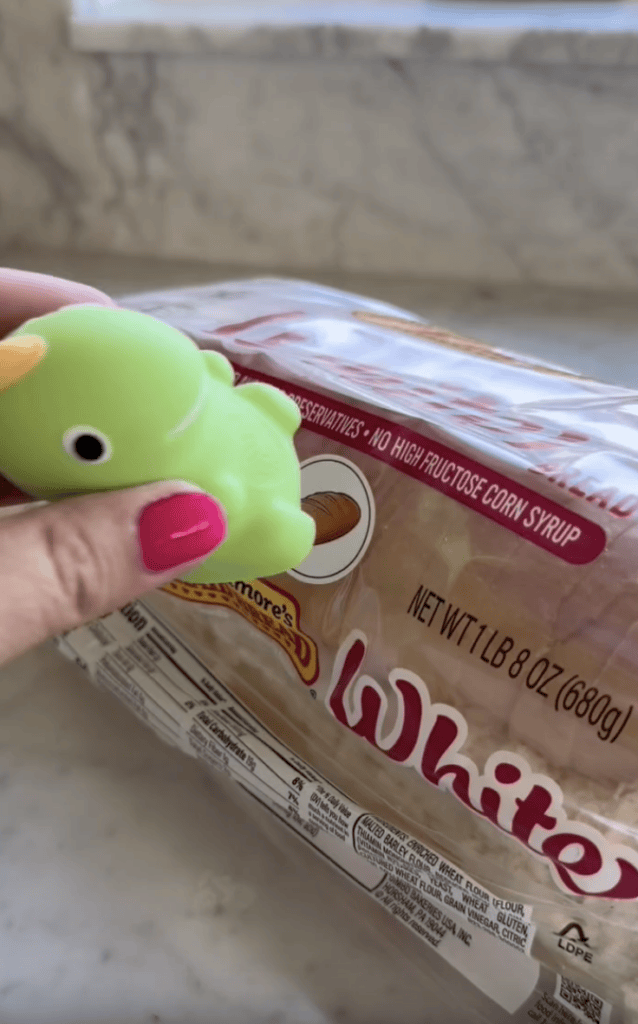 A hand with pink nail polish holds a small green toy bird above a loaf of sliced white bread in plastic packaging on a kitchen counter.
