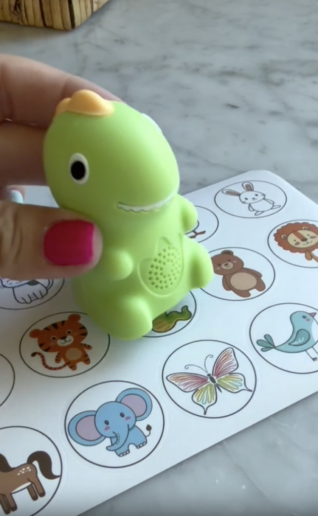 A hand with pink nail polish holds a small green dinosaur toy above a sheet featuring illustrated animals, including an elephant, butterfly, tiger, bird, and lion on a marble surface.