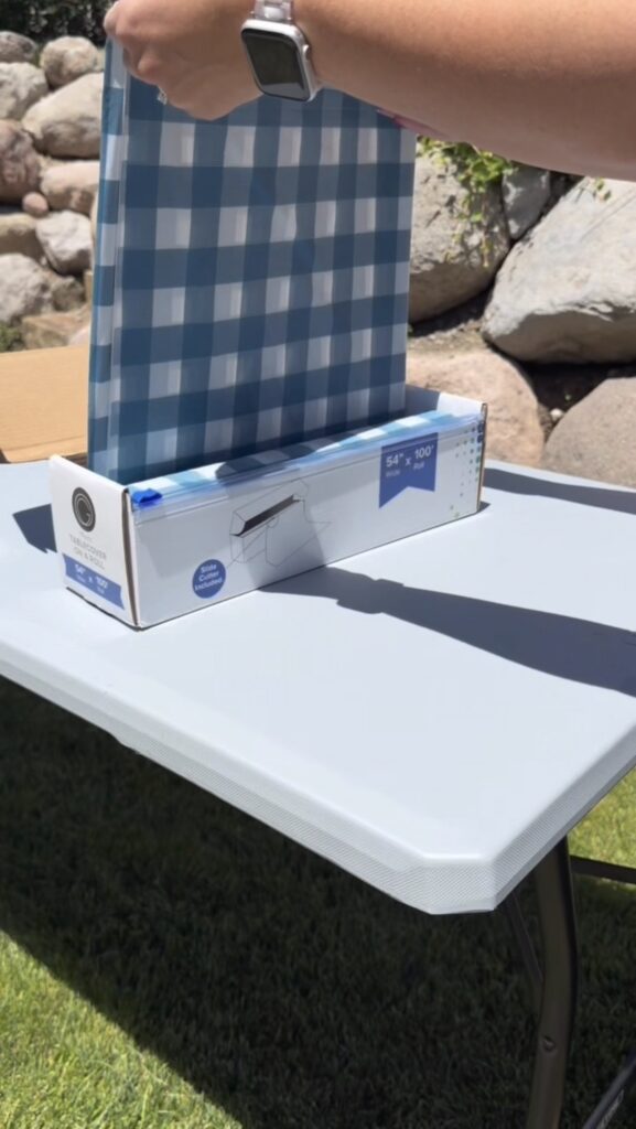 A person pulls a blue and white checkered table cover from a box dispenser onto a white folding table outdoors near rocks.