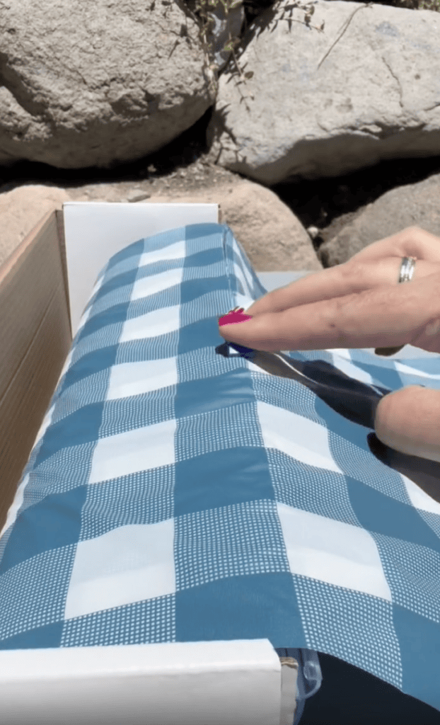 A hand with pink nails and a ring smooths a blue and white checkered tablecloth over a table outdoors, with rocks visible in the background.
