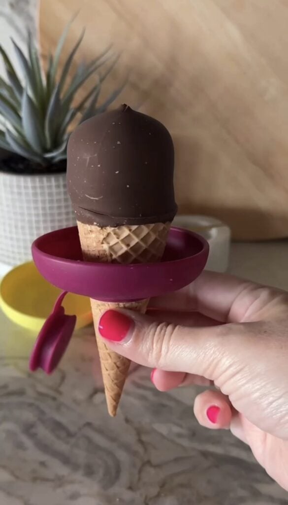 A hand with pink nail polish holds an ice cream cone with a chocolate coating and a pink drip catcher ring around the cone. A potted plant and yellow container are in the blurred background.