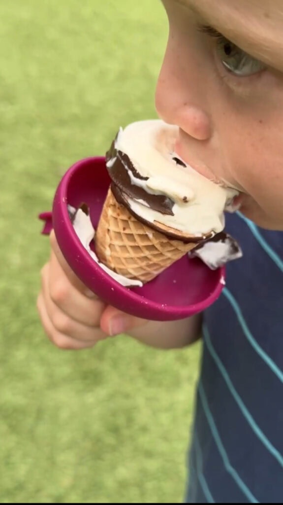 A child wearing a blue striped shirt eats a chocolate-dipped ice cream cone using a pink spill guard, with green grass in the background.