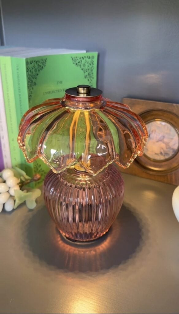 A pink and orange ribbed glass lamp sits on a shelf beside green books, a small grape ornament, and a framed picture.