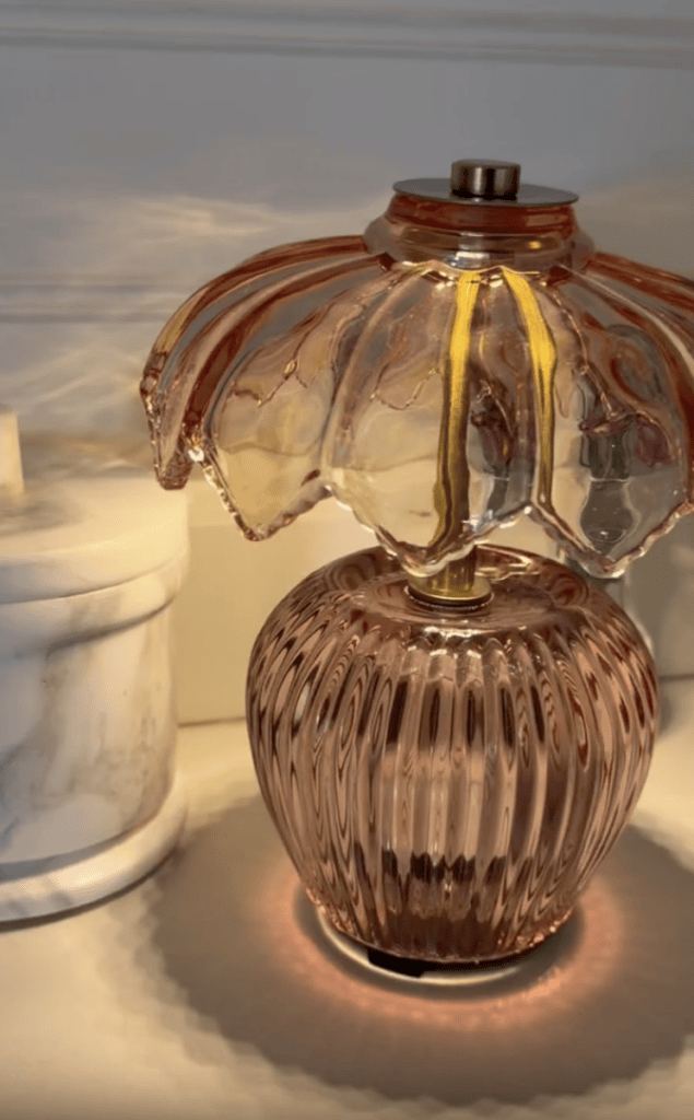 A pink glass lamp with a fluted, bulbous base and a flower-shaped shade sits on a surface next to a white marble container. Warm light creates soft reflections on the wall and surface.