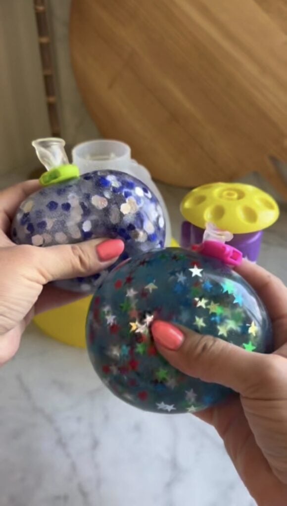 Two hands hold colorful sensory stress balls filled with liquid, glitter, and star-shaped confetti. The balls are tied with balloon knots. A yellow and purple container and a wooden board are visible in the blurred background.