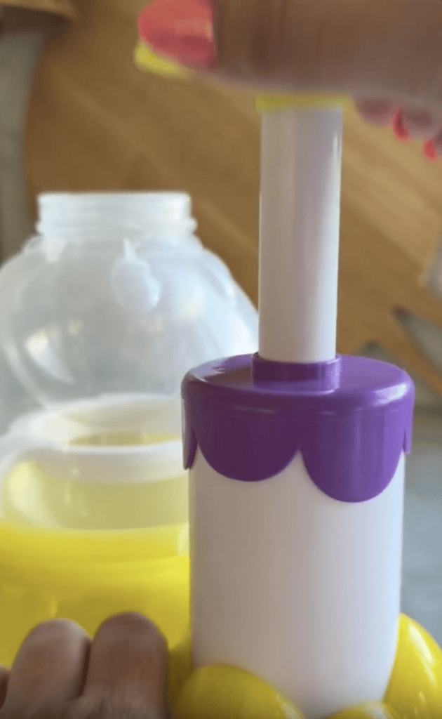 A hand presses a white handle with a purple top onto a yellow container, with an open bubble solution bottle in the background.