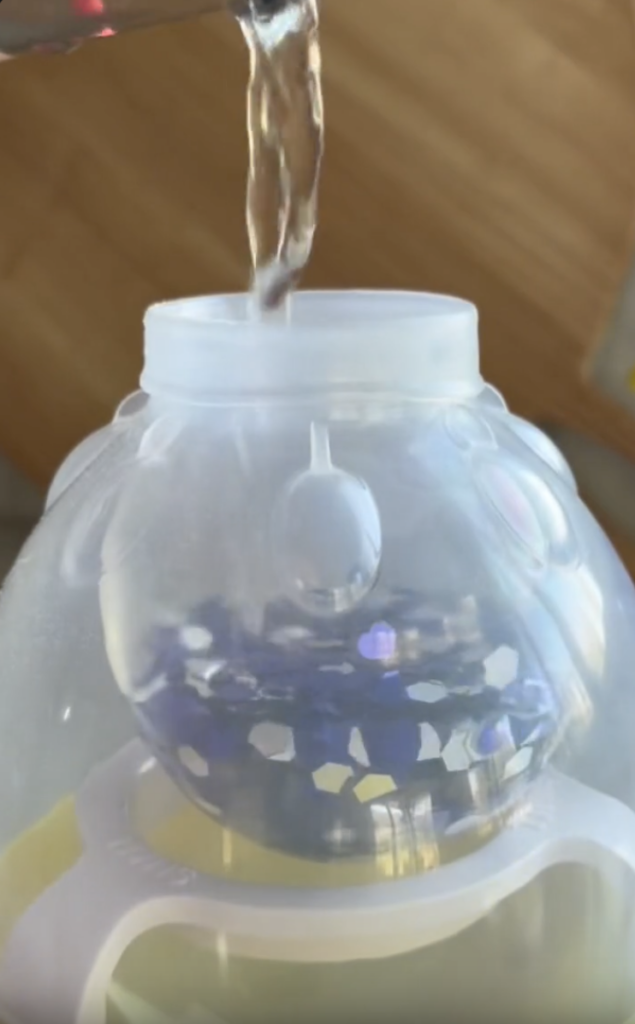Water is being poured into a translucent container filled with blue and white decorative beads or crystals, seen through the open top.