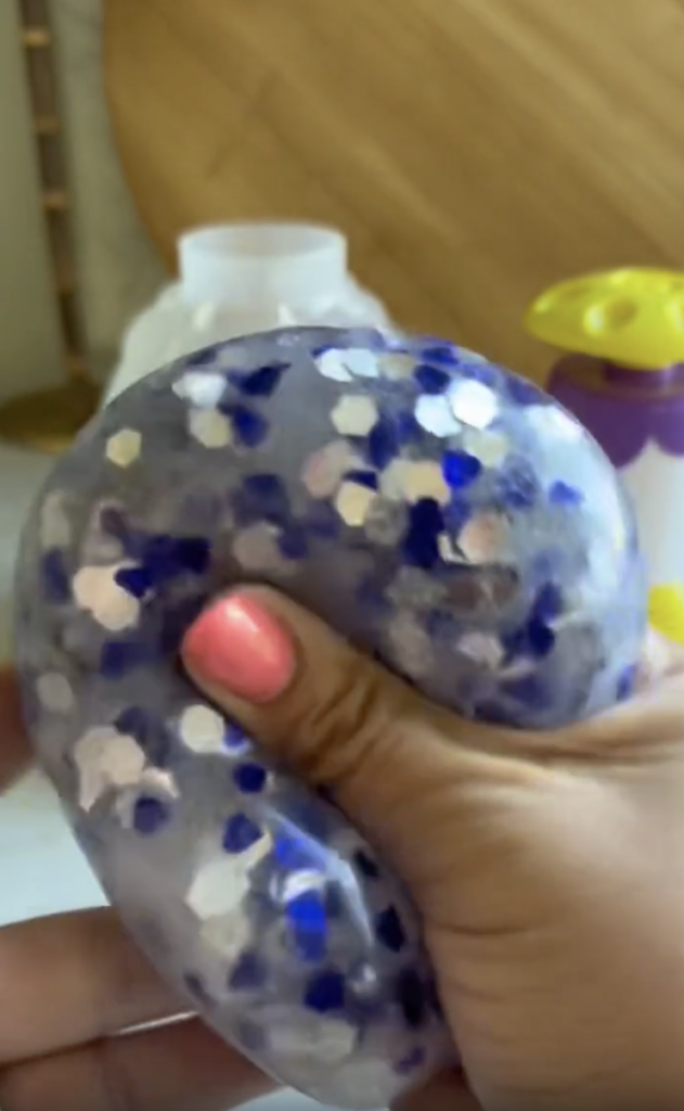 A hand with pink nail polish squeezes a translucent, glittery slime with blue and white hexagon shapes. Bottles and a wooden board are blurred in the background.