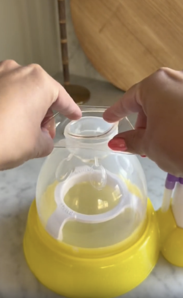 Two hands hold a clear lid above a yellow egg cooker with a transparent dome, placed on a marble countertop. A wooden board and lamp are in the blurred background.