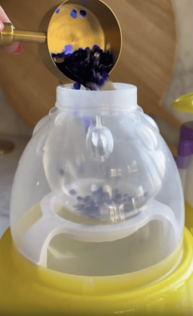 A gold measuring cup pours blue and black beads into a translucent container placed on a yellow base, with a wooden board and other kitchen items in the background.