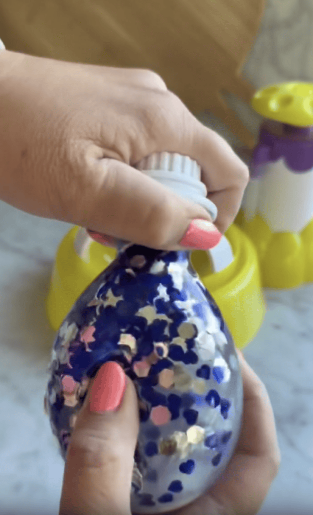 A hand with pink nail polish holds a blue balloon filled with star-shaped confetti, while twisting a cap onto its neck. Yellow objects are blurred in the background.